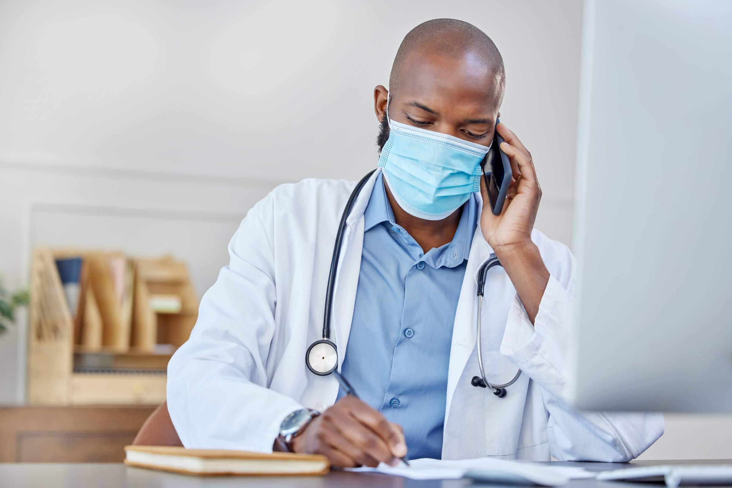 Shot of a young doctor on a call while writing in a notebook at work